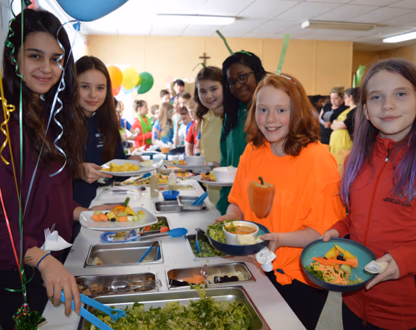 Group of smiling children holding plates of food in a buffet line with colorful balloons in background.