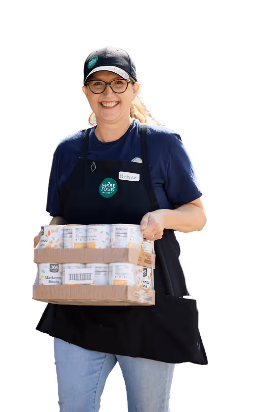 Smiling Whole Foods worker named Nichole holding a box of canned goods wearing apron and cap.