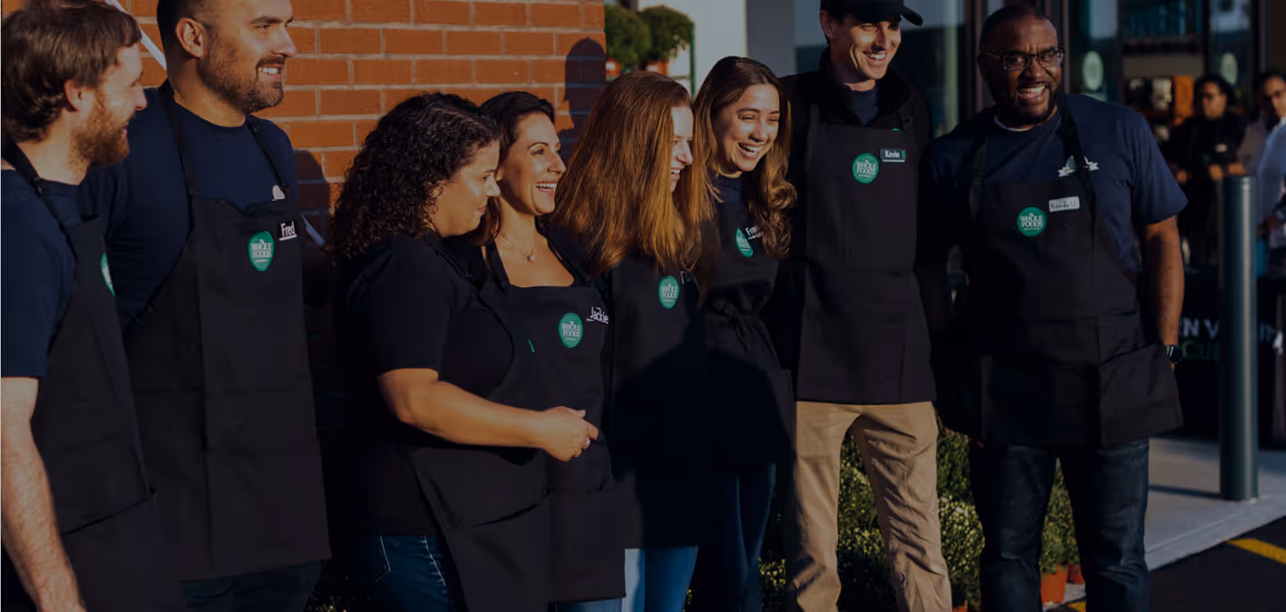 Seven people wearing black aprons posing side-by-side outdoors in front of a brick wall.