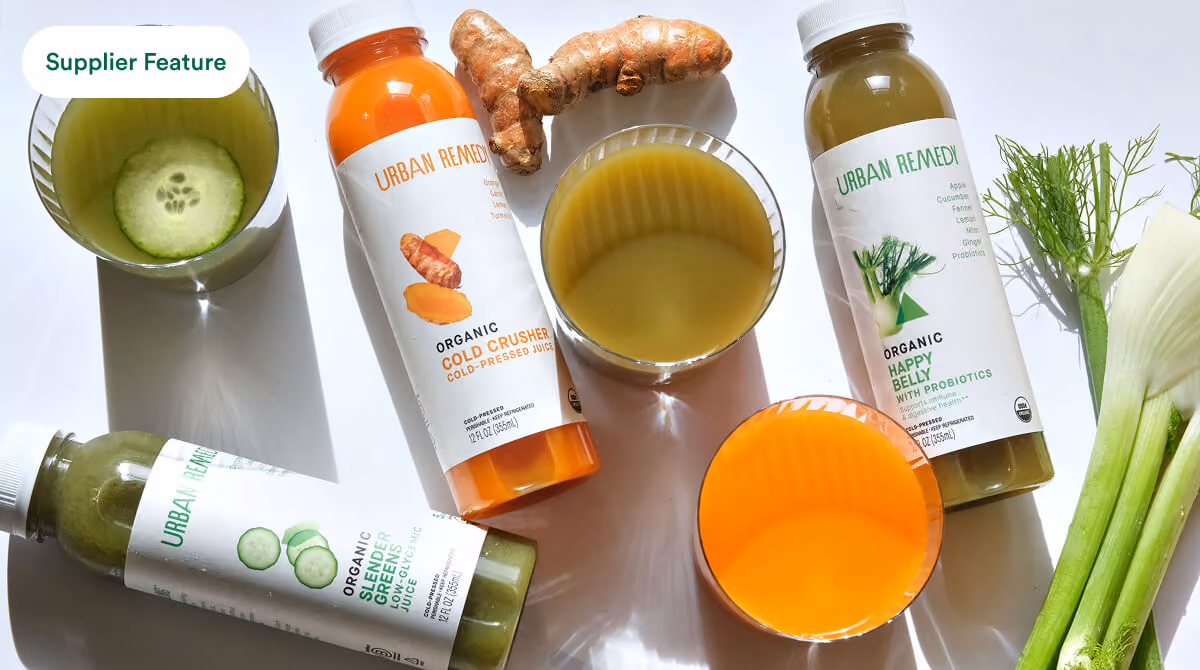 Three Urban Remedy organic juice bottles with glasses and fresh ingredients like turmeric and fennel.