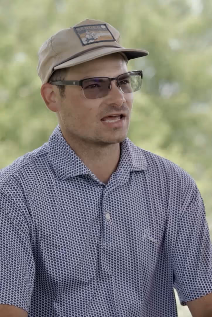 Man wearing patterned polo shirt, glasses, and beige cap is speaking outdoors with blurred greenery behind.