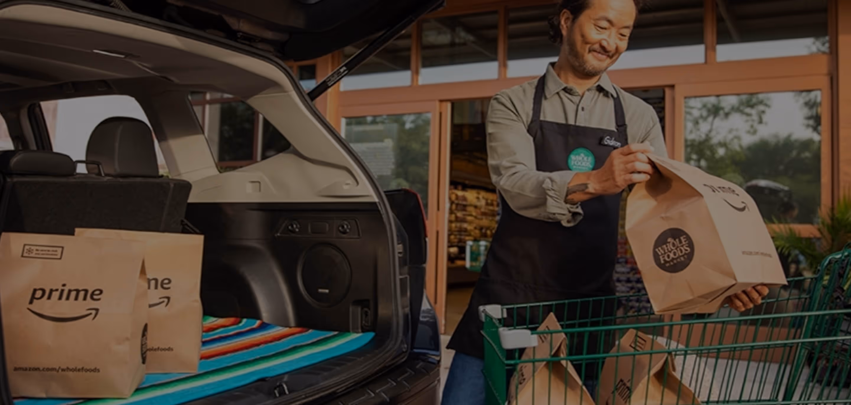 Man in apron loading Whole Foods Prime grocery bags into an open car trunk outside a store.