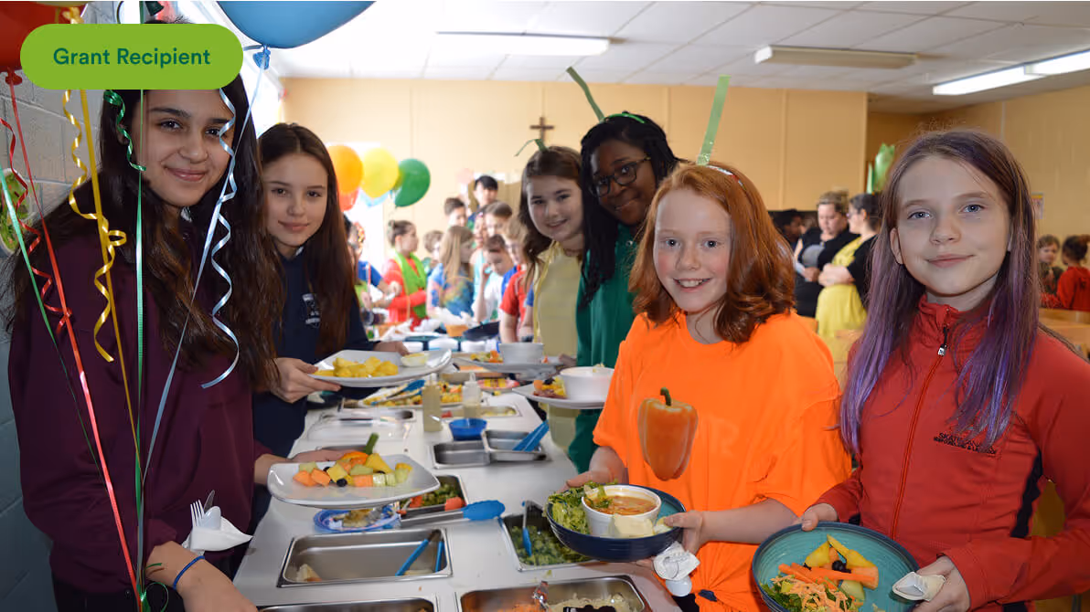 Group of children smiling while holding plates at a cafeteria food serving line with balloons nearby.