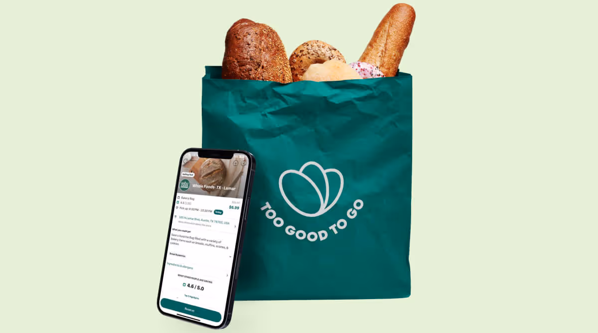 Green bag labeled Too Good To Go filled with assorted bread and a phone showing the app interface.