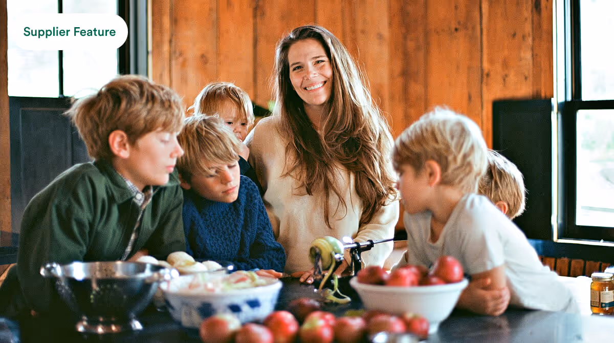 Woman with five children peeling apples at a kitchen table with bowls of fruit and a window background.